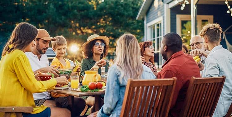 A group of people of various ages sit around a wooden outdoor table, enjoying a meal together. The table has food, drinks, and fresh produce. String lights and greenery create a warm, festive atmosphere.